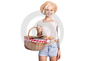 Young blonde woman with tattoo wearing summer hat and holding picnic wicker basket with bread looking positive and happy standing