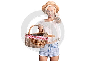 Young blonde woman with tattoo wearing summer hat and holding picnic wicker basket with bread pointing with finger to the camera