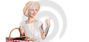 Young blonde woman with tattoo wearing summer hat and holding picnic wicker basket with bread pointing thumb up to the side