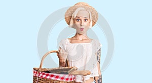 Young blonde woman with tattoo wearing summer hat and holding picnic wicker basket with bread scared and amazed with open mouth