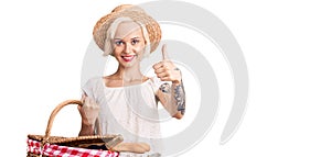 Young blonde woman with tattoo wearing summer hat and holding picnic wicker basket with bread smiling happy and positive, thumb up