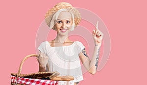 Young blonde woman with tattoo wearing summer hat and holding picnic wicker basket with bread surprised with an idea or question