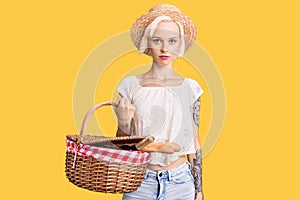 Young blonde woman with tattoo wearing summer hat and holding picnic wicker basket with bread thinking attitude and sober