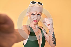 Young blonde woman with tattoo wearing swimwear taking a selfie annoyed and frustrated shouting with anger, yelling crazy with