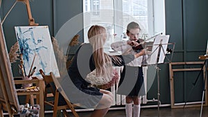 Young blonde woman teacher teaching a little girl student how to properly hold a bow while playing violin