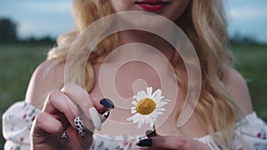 A young blonde woman tears off the petals of a daisy standing in a green field