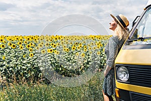 Young blonde woman travelling by campervan. Overlooking sunflower field on sunny summer day