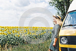 Young blonde woman travelling by campervan. Overlooking sunflower field on sunny summer day