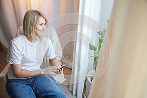 Young blonde woman using cell phone at home. Girl is resting, chating and looking in camera on smartphone near window at