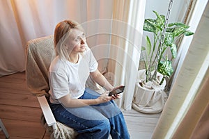 Young blonde woman using cell phone at home. Girl is resting, chating and looking in camera on smartphone near window at