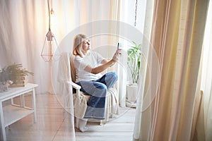 Young blonde woman using cell phone at home. Girl is resting, chating and looking in camera on smartphone near window at