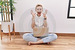 Young blonde woman using computer laptop sitting on the floor at the living room celebrating crazy and amazed for success with