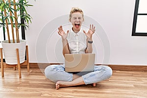 Young blonde woman using computer laptop sitting on the floor at the living room celebrating mad and crazy for success with arms