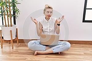 Young blonde woman using computer laptop sitting on the floor at the living room clueless and confused expression with arms and