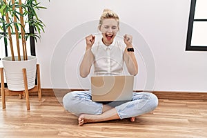Young blonde woman using computer laptop sitting on the floor at the living room excited for success with arms raised and eyes
