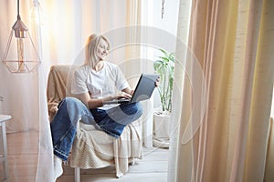 Young blonde woman using laptop computer at home. Girl is resting, chating and looking in camera near window at home
