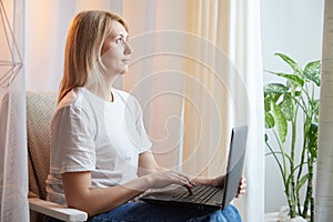 Young blonde woman using laptop computer at home. Girl is resting, chating and looking in camera near window at home