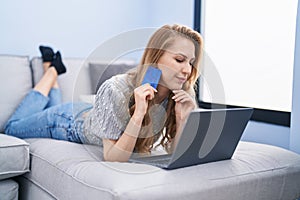 Young blonde woman using laptop and credit card lying on sofa at home