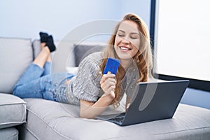 Young blonde woman using laptop and credit card lying on sofa at home