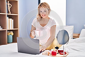 Young blonde woman using laptop drinking coffee at bedroom