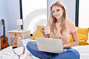 Young blonde woman using laptop drinking coffee sitting on bed at bedroom