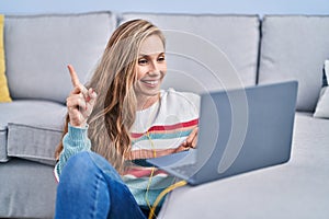 Young blonde woman using laptop at home smiling happy pointing with hand and finger to the side