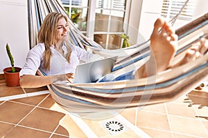 Young blonde woman using laptop lying on hammock at terrace