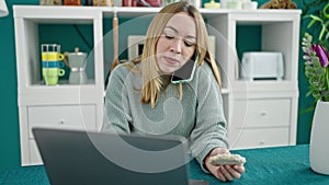 Young blonde woman using laptop talking on smartphone eating rice cake at dinning room