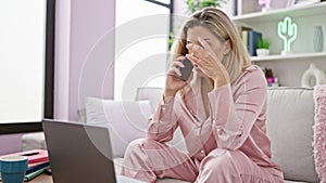 Young blonde woman using laptop talking on smartphone with serious face at home