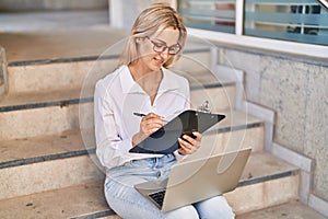 Young blonde woman using laptop and write on clipboard at street