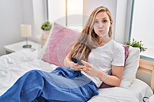 Young blonde woman using smartphone on bed puffing cheeks with funny face
