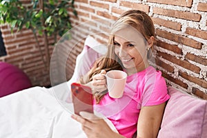 Young blonde woman using smartphone drinking coffee at bedroom