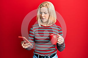 Young blonde woman using smartphone and drinking a cup of coffee clueless and confused expression