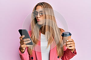 Young blonde woman using smartphone and drinking a cup of coffee looking at the camera blowing a kiss being lovely and sexy