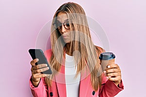 Young blonde woman using smartphone and drinking a cup of coffee making fish face with mouth and squinting eyes, crazy and comical