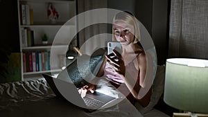 Young blonde woman using smartphone and laptop sitting on bed at bedroom