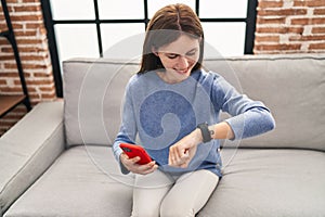 Young blonde woman using smartphone looking watch at home