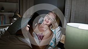 Young blonde woman using smartphone lying on bed smiling at bedroom