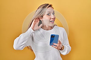 Young blonde woman using smartphone typing message smiling with hand over ear listening an hearing to rumor or gossip