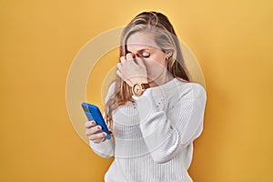 Young blonde woman using smartphone typing message tired rubbing nose and eyes feeling fatigue and headache