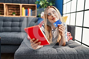 Young blonde woman using touchpad and credit card lying on sofa at home