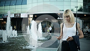 Young blonde woman walks in the city passes near the fountain, wets her hands, touches the water.