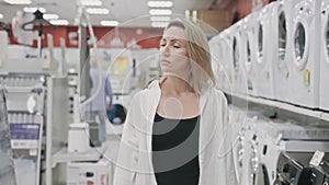 A young blonde woman walks through an electronics hypermarket and looks at washing machines. A woman is choosing a