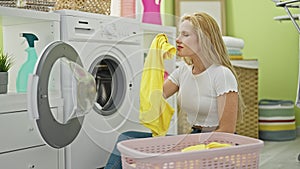 Young blonde woman washing clothes smelling dirty t shirt at laundry room