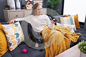 Young blonde woman watching movie sitting on sofa with dog at home