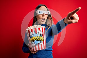 Young blonde woman wearing 3d glasses and eating pack of popcorn watching a movie on cinema Pointing with finger surprised ahead,