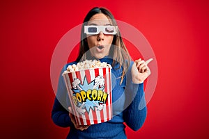 Young blonde woman wearing 3d glasses and eating pack of popcorn watching a movie on cinema Surprised pointing with finger to the