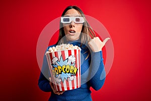 Young blonde woman wearing 3d glasses and eating pack of popcorn watching a movie on cinema Surprised pointing with hand finger to
