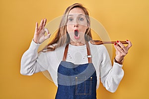 Young blonde woman wearing apron tasting food holding wooden spoon afraid and shocked with surprise and amazed expression, fear