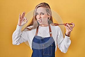 Young blonde woman wearing apron tasting food holding wooden spoon clueless and confused expression